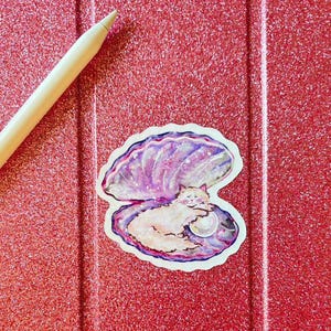 May include: A sticker depicting a white cat nestled in a purple and blue seashell, with a pearl. The sticker is on a red glittery surface, with a light-coloured stylus in the upper left corner.