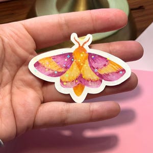 May include: A colorful moth sticker with a white border. The moth has a vibrant design with orange, yellow, and purple wings, and a small antenna. The sticker is held in a hand.
