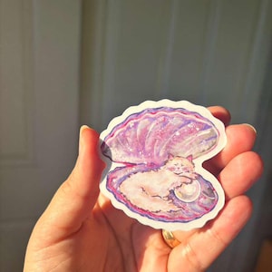 May include: A sticker depicting a cat inside a pearl oyster shell. The shell is painted in shades of pink and purple, with a pearl next to the cat. The cat is light-coloured, creating a charming design.