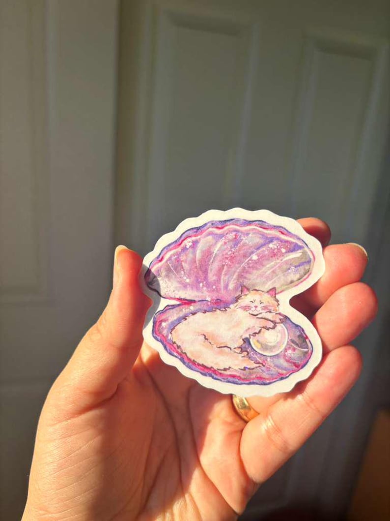 May include: A sticker depicting a cat inside a pearl oyster shell. The shell is painted in shades of pink and purple, with a pearl next to the cat. The cat is light-coloured, creating a charming design.