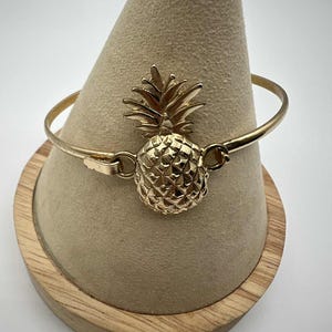 Puede incluir: Pulsera de brazalete dorada con un dije de piña detallado. El dije de piña es texturizado y tiene una corona de hojas. La pulsera se exhibe en un soporte cónico beige con base de madera. Joyería.