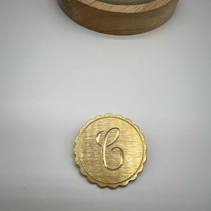 May include: A gold-colored, round monogram with a scalloped edge. The letter "C" is elegantly scripted in the center. The monogram is set against a white background, with a wooden base visible in the upper portion.