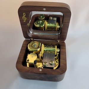 May include: A wooden music box with a hinged lid and a mirrored interior. The box is open, revealing the gold-colored mechanical components. A gold treble clef is on the top left corner of the lid. The box is made of dark brown wood.