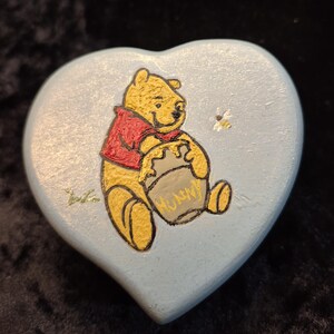 May include: A heart-shaped, light blue trinket box featuring a hand-painted image of Winnie the Pooh holding a honey pot. The bear is yellow with a red shirt. A small bee is painted near the bear. The word "HONEY" is on the honey pot.
