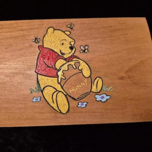 May include: Wooden box with a hand-painted illustration of Winnie the Pooh. The yellow bear is wearing a red shirt and holding a honey pot labeled "Hunny". Bees and flowers are also painted on the wooden surface.