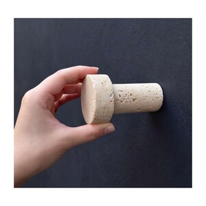 Puede incluir: Un gancho de pared cilíndrico hecho de piedra porosa de color beige claro con una cara frontal circular plana y una textura uniforme. El gancho presenta un acabado superficial natural y poroso característico del travertino en tono neutro.