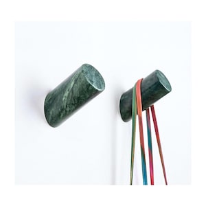May include: Two cylindrical wall hooks crafted from dark green marble with natural veining. One hook is empty, while the other holds a set of thin, multicoloured leather straps that hang vertically from the polished stone surface.