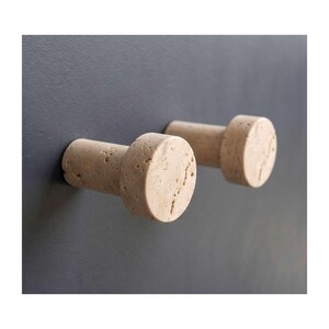 Può includere: Due ganci da parete cilindrici realizzati in pietra travertino beige con una superficie porosa e strutturata. Ogni gancio presenta una faccia anteriore piatta e circolare e uno stelo robusto che si estende dal centro, montato orizzontalmente.