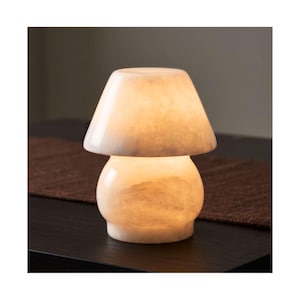 Może przedstawiać: Lampa stołowa w kształcie grzyba wykonana z półprzezroczystego białego kamienia z naturalnym żyłkowaniem. Lampa posiada zaokrągloną podstawę oraz szeroki, rozkloszowany klosz, który po włączeniu emituje miękkie, ciepłe światło.