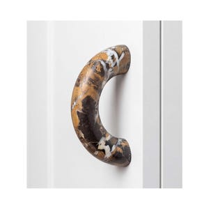 May include: A curved cabinet handle featuring a polished stone finish with a marbled pattern in shades of dark brown, tan, and white. The handle is mounted onto a vertical white panel, showcasing a smooth, rounded arch shape and a glossy texture.