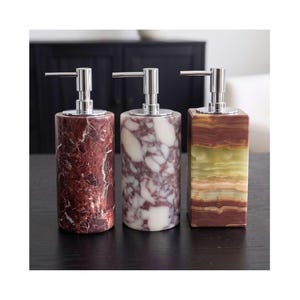 Può includere: Tre dispenser per sapone con erogatori in metallo argentato. Due dispenser sono cilindrici con motivi in marmo rosso scuro e bianco. Il terzo dispenser è rettangolare con fasce orizzontali di texture in pietra verde e marrone traslucida.