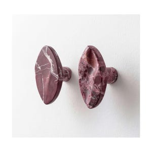 Può includere: Due pomelli per mobili di forma ovale realizzati in marmo color bordeaux intenso con venature bianche distinte. Ogni pomello presenta una superficie liscia e lucida e una base cilindrica, che mette in risalto la texture naturale della pietra.