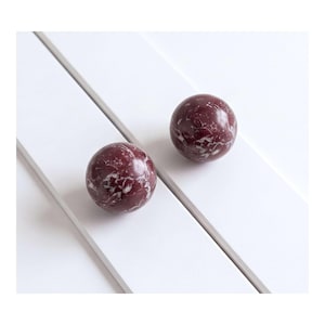 May include: Two spherical objects made of deep burgundy stone with prominent white veining throughout the surface. The spheres have a smooth, polished finish and are positioned separately, showcasing the natural marble-like pattern of the stone.