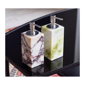 Può includere: Due dispenser di sapone rettangolari con erogatori cromati. Uno presenta marmo bianco con venature viola scuro, mentre l'altro è realizzato in pietra traslucida verde chiaro con motivi minerali naturali e variazioni cromatiche.
