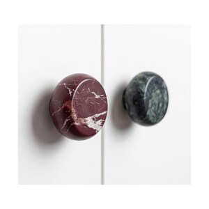 Peut inclure: Deux boutons de placard ronds montés sur une surface blanche. Le bouton de gauche est en marbre bordeaux profond avec des veines blanches, et le bouton de droite est en marbre vert forêt foncé avec des veines subtiles sur la surface polie.