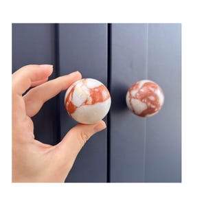 Puede incluir: Un pomo de armario esférico con un patrón de piedra veteada en blanco y naranja rojizo pulido. El pomo está montado en una puerta de armario azul oscuro, con un segundo pomo idéntico visible al fondo, mostrando un acabado liso y brillante.