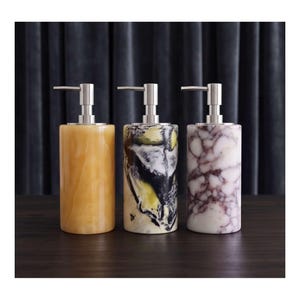 May include: Three cylindrical soap dispensers with silver-toned metal pump mechanisms. The first dispenser is a solid translucent amber colour, the second features a marbled pattern of black, white, and yellow, and the third has a white and purple marble design.