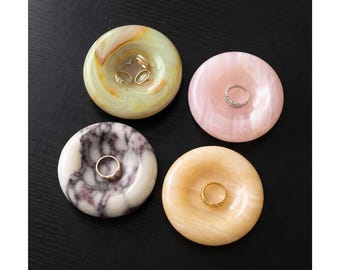 May include: Four circular stone ring dishes arranged in a square formation. One dish is pale green with brown swirls, one is solid light pink, one is white with dark purple veining, and one is solid light orange. Each dish holds a gold or silver ring.