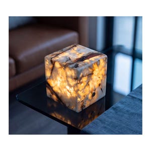 Pode incluir: Uma lâmpada em forma de cubo feita de pedra branca translúcida com veios castanhos escuros. A pedra emite uma luz quente a partir do interior, realçando os padrões e texturas naturais do material. Um pequeno interruptor metálico circular é visível.