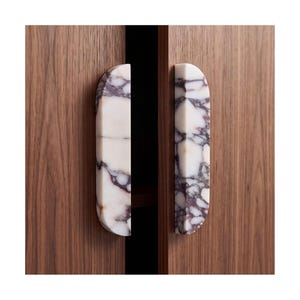 May include: Two vertical semi-circular cabinet handles made of white marble with prominent dark purple veining. The handles are mounted on two adjacent dark brown wooden cabinet doors with a visible vertical wood grain pattern.