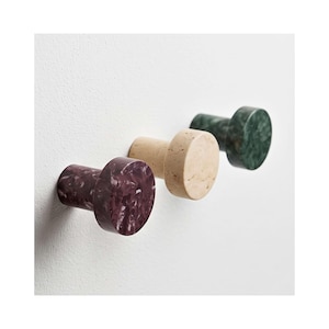 May include: Three cylindrical wall hooks with flat circular faces. The front hook is a deep purple with white flecks, the middle hook is a light beige with a porous texture, and the back hook is a dark green with a marbled pattern.