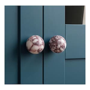 May include: Two spherical cabinet knobs featuring a polished white and deep purple marble pattern. The knobs are mounted on a flat, matte teal blue cabinet door surface with vertical paneling details, creating a distinct contrast in texture.