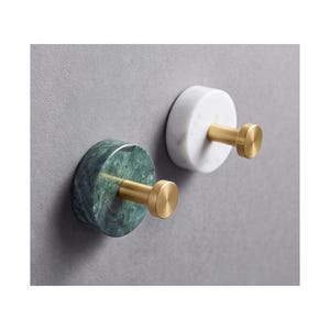 May include: Two wall hooks featuring circular bases and cylindrical pegs. One hook has a dark green marble base with a brushed gold finish peg, while the other has a white marble base with a matching brushed gold finish peg.