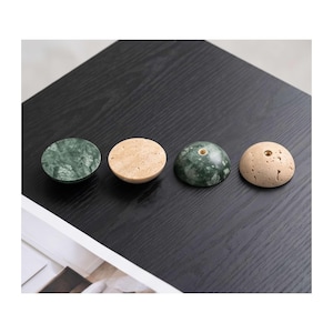 May include: Four hemispherical knobs arranged in a row. Two are made of dark green marble with white veining, and two are made of beige porous stone. Each knob features a small, centered brass-colored metal insert on the top surface.
