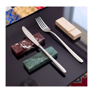 Puede incluir: Un cuchillo y un tenedor de metal plateado apoyados sobre tres soportes rectangulares de piedra. Los soportes están hechos de mármol pulido en rojo oscuro con vetas blancas, verde oscuro con patrones sutiles y una piedra de color beige claro.
