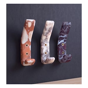 May include: Three wall-mounted hooks made of polished stone with distinct marble patterns. The left hook is reddish-brown, the center is white and grey, and the right is dark purple with white veining. Each hook features two brass-lined screw holes.