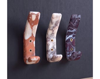 May include: Three wall-mounted hooks made of polished stone with distinct marble patterns. The left hook is reddish-brown, the center is white and grey, and the right is dark purple with white veining. Each hook features two brass-lined screw holes.