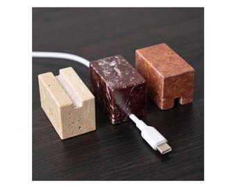 May include: Three rectangular stone cable organizers in beige, dark maroon, and reddish-brown. Each block features a narrow vertical slot on the top surface designed to hold a white charging cable, with one cable currently threaded through the center block.
