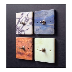 May include: Four square light switches with toggle levers. The top left is white with purple veining, the top right is blue with white swirls, the bottom left is orange-brown with a wood-like grain, and the bottom right is a solid pale green.