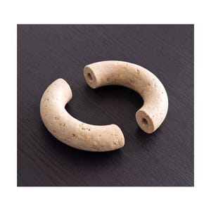Peut inclure: Deux poignées de meuble incurvées en forme de demi-cercle avec une finition beige texturée rappelant la pierre. Chaque pièce présente une forme cylindrique avec une surface poreuse et un trou de montage circulaire visible à chaque extrémité.