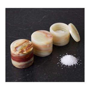 May include: Two cylindrical stone containers with lids, one featuring deep red and brown banding and the other with pale cream and translucent green layers. A small pile of white salt sits next to the open container, which also contains salt.