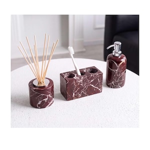 May include: A bathroom accessory set featuring a dark red marble finish with white veining. The set includes a cylindrical reed diffuser with wooden sticks, a rectangular toothbrush holder with three circular openings, and a soap dispenser.