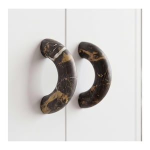 May include: Two semi-circular cabinet pulls featuring a dark brown and black base with prominent gold and white veining patterns. These curved handles are mounted onto a plain white surface, showcasing a smooth, polished finish throughout.