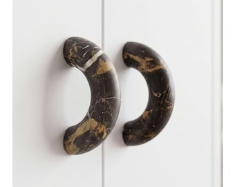 May include: Two semi-circular cabinet pulls featuring a dark brown and black base with prominent gold and white veining patterns. These curved handles are mounted onto a plain white surface, showcasing a smooth, polished finish throughout.