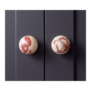 May include: Two spherical cabinet knobs made of polished stone with a cream-colored base and deep red, marbled veining. The knobs are mounted on a dark charcoal gray cabinet door with vertical paneling, showcasing a smooth, rounded surface.