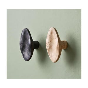 Puede incluir: Dos ganchos de pared con forma ovalada y orgánica, con una superficie porosa y texturizada. Un gancho es de color gris carbón oscuro y el otro es de color beige claro. Cada gancho tiene una base cilíndrica que sobresale de la pared.
