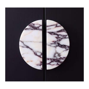 May include: Two semi-circular cabinet pulls made of white marble with prominent dark purple and black veining. The two pieces are positioned vertically against a solid black surface, forming a complete circle when placed side by side.
