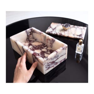 May include: A rectangular white and purple marble box with a matching lid featuring a dark purple crystal handle. The marble has a distinct veined pattern throughout the stone. A small clear glass perfume bottle with a gold cap sits nearby.