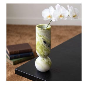 May include: A cylindrical stone vase with a spherical base, featuring a marbled pattern of white and various shades of green. A single stem with three white orchid flowers is placed inside the opening of the vase, extending upwards.