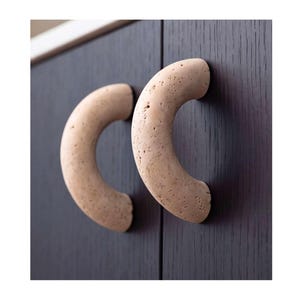 Puede incluir: Dos tiradores de armario semicirculares con una textura porosa, de color beige claro, similar a la piedra. Estos tiradores curvos están montados verticalmente en una puerta de armario de madera marrón oscuro, con una superficie picada.