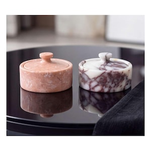 Puede incluir: Dos recipientes redondos de piedra con tapa, cada uno con un pequeño pomo. Uno es de color rosa claro sólido con vetas blancas sutiles, mientras que el otro es blanco con vetas de mármol prominentes en tonos púrpura oscuro y granate.
