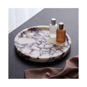 Könnte beinhalten: Ein rundes Marmortablett mit weißer Basis und dunkelvioletter Maserung. Auf der Oberfläche stehen zwei kleine Glasflaschen mit Metallverschlüssen, eine mattiert und eine bernsteinfarben. Das Tablett hat einen erhöhten Rand.