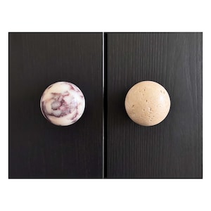 Può includere: Due pomelli sferici per armadi montati su ante in legno scuro. Il pomello sinistro è bianco con venature di marmo viola scuro e una finitura liscia. Il pomello destro è di colore beige chiaro con una superficie porosa, ruvida e irregolare.