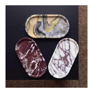 May include: Three oval-shaped trays with smooth, raised edges. One tray features a yellow, grey, and white marbled pattern. Another tray displays a deep burgundy and white veined design. The third tray has a white base with dark red veining.