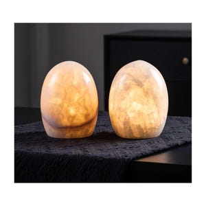 May include: Two egg-shaped lamps made of translucent, pale stone with a crystalline, marbled texture. Each lamp emits a warm, soft internal glow, highlighting the natural variations and jagged patterns within the stone structure.