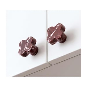 May include: Two clover-shaped cabinet knobs made of dark red marble with white veining. Each knob features a smooth, polished surface and is mounted onto a flat, white cabinet door panel, providing a distinct contrast in color and texture.